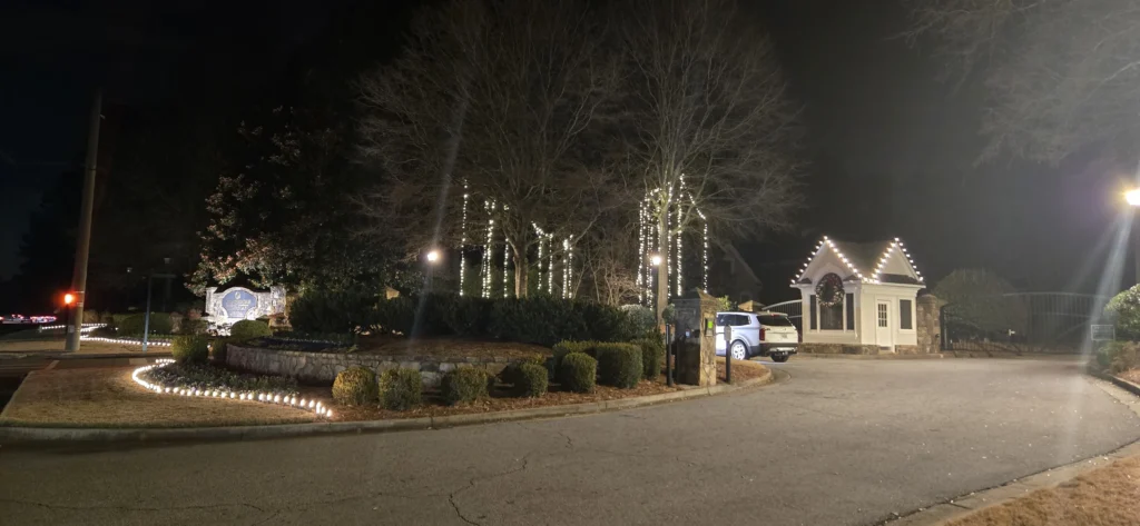 Wide nighttime view of an entrance with warm white landscape lights, wrapped trees, and a gatehouse